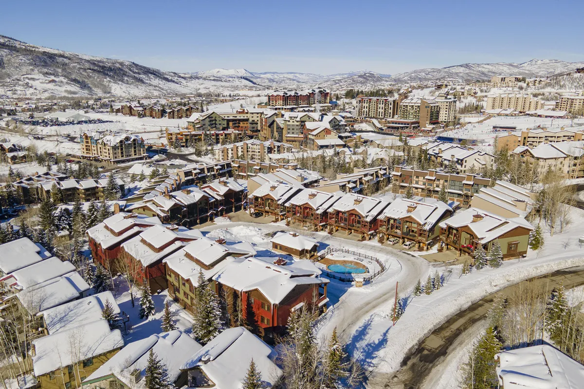 An inviting exterior scene featuring snow-covered roofs, a centrally located swimming pool, and sweeping mountain views in the background. The cluster of rustic buildings creates a picturesque alpine village vibe, while the pool and mountains emphasize year-round relaxation—whether after skiing in winter or hiking in summer.
