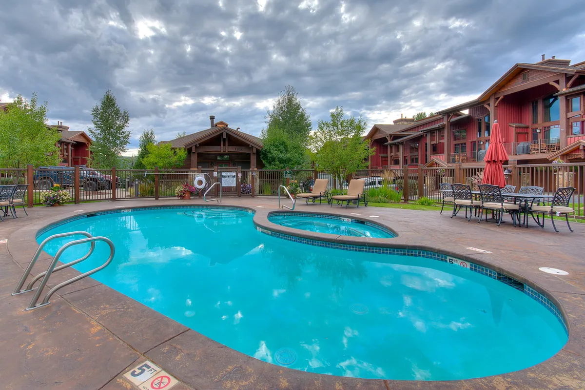 This exterior image showcases a sparkling swimming pool and a bubbling hot tub surrounded by lounge chairs, with a red umbrella adding a pop of color. Lush greenery and trees frame the area, creating a tranquil, resort-style atmosphere. The setup is ideal for socializing or relaxing, echoing the section's emphasis on resort amenities and year-round enjoyment.