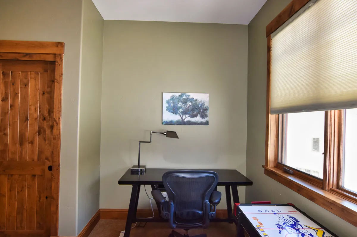 A well-lit bedroom workspace featuring a desk, an ergonomic office chair, and a painting above the workspace. Natural light filters in through the window, creating a serene setting for working or studying. The decor is simple yet focused, supporting productivity.