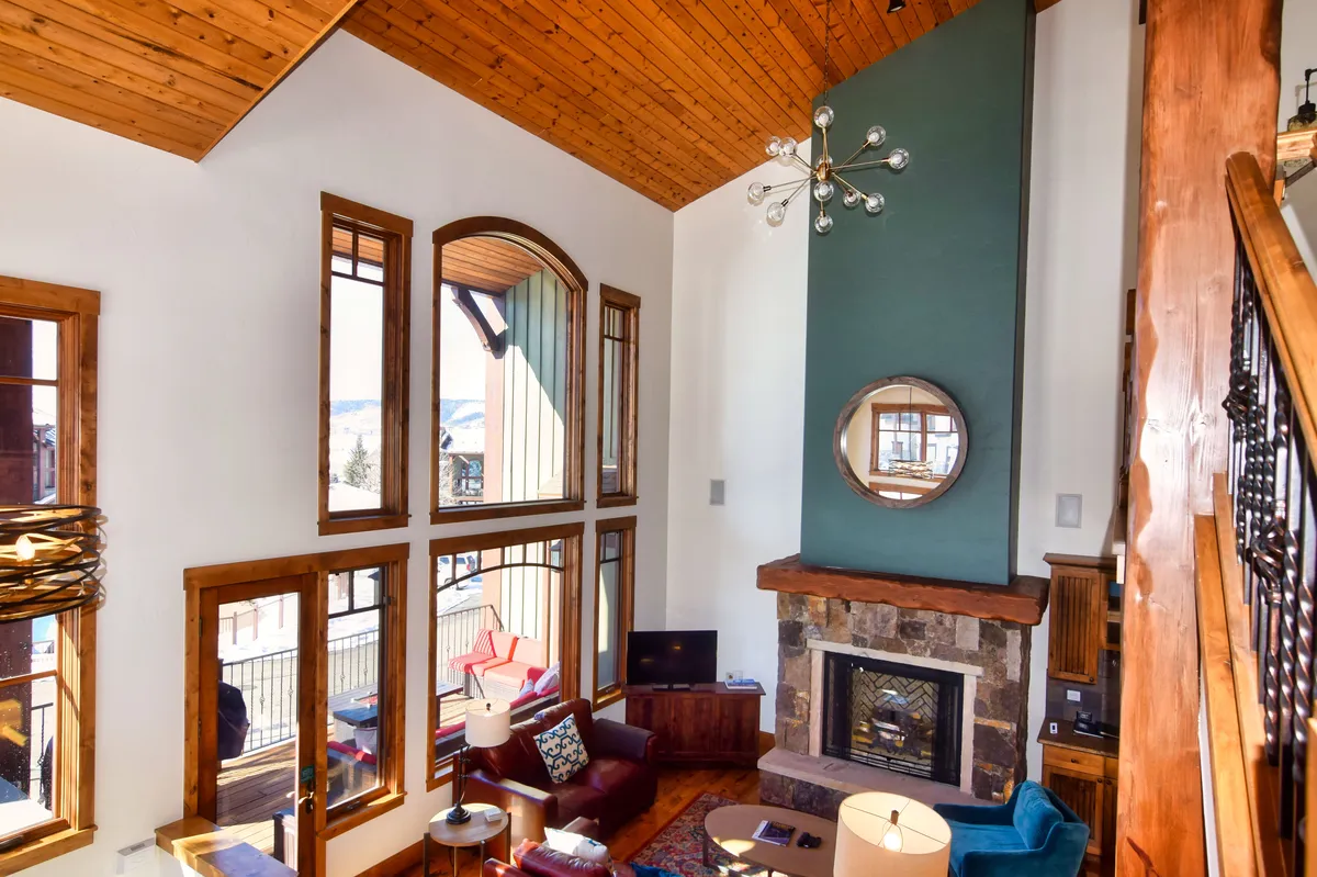 This image showcases a spacious living area filled with natural light from large windows, a central fireplace, a striking chandelier, and exposed wooden beams. The view through the windows hints at the surrounding scenery, subtly connecting the interior space to the outdoors.