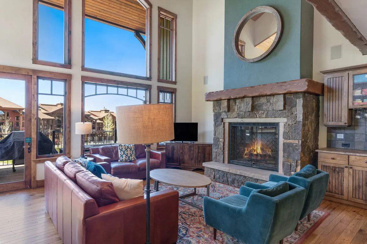 The hero image showcases a spacious, luxuriously furnished living room with soaring ceilings, a grand stone fireplace as the centerpiece, expansive floor-to-ceiling windows revealing mountain views, and plush seating arranged for cozy gatherings. Warm lighting from a decorative lamp and natural sunlight create an inviting, serene atmosphere perfect for relaxation and socializing.