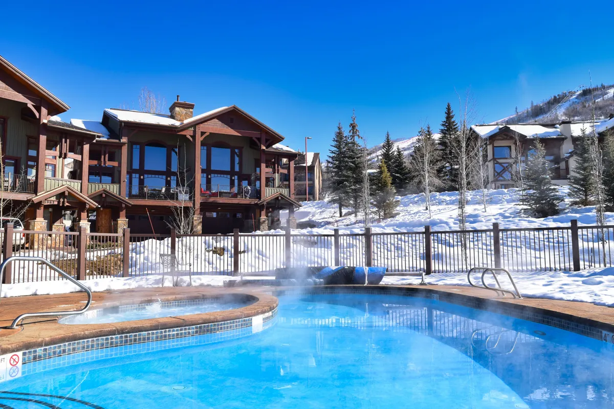 An outdoor hot tub set in a snow-covered landscape, bordered by trees and the building's facade. The scene evokes a serene and cozy winter escape, perfect for unwinding after a day on the slopes.