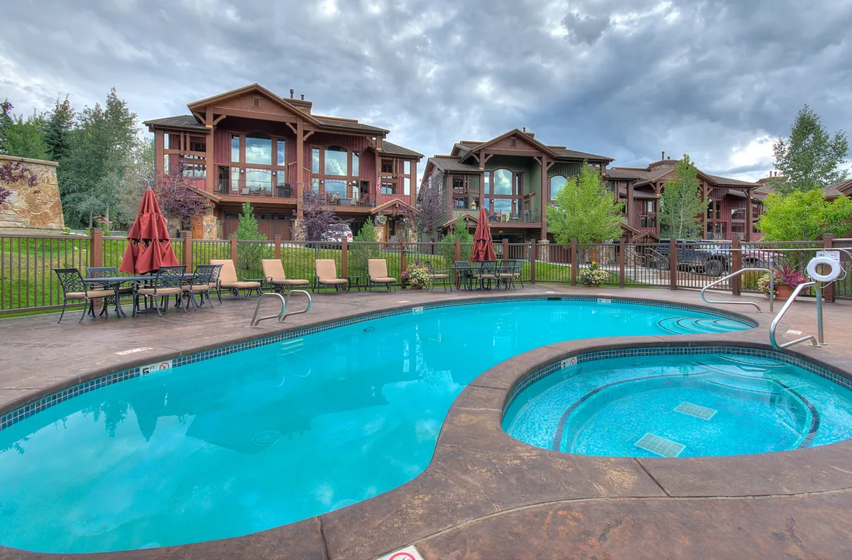 A bright, inviting community pool area featuring a heated swimming pool and adjacent hot tub surrounded by comfortable lounge chairs and patio umbrellas. Lush greenery frames the space, evoking a relaxing, resort-like ambiance.