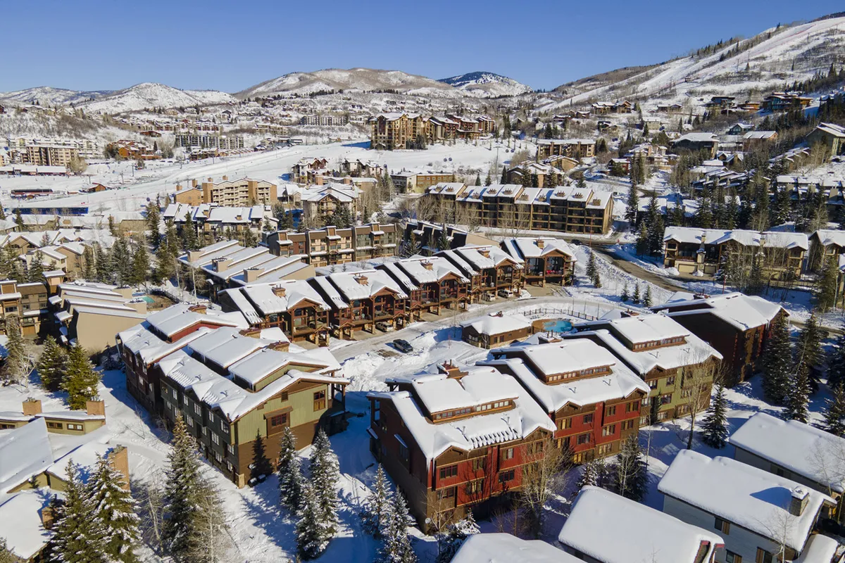 A scenic exterior view featuring snow-covered rooftops, pine trees, ski slopes, and surrounding residential buildings. The image conveys a cozy, adventurous atmosphere and highlights the proximity to Steamboat's iconic outdoor activities and community life.