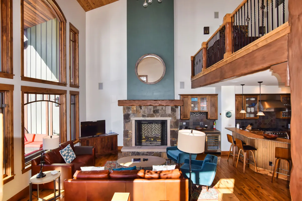 A spacious open-concept living area featuring a large stone fireplace flanked by plush seating, high vaulted ceilings, and expansive windows that flood the room with natural light. The kitchen area, visible in the background, boasts modern finishes and an inviting layout ideal for entertaining.