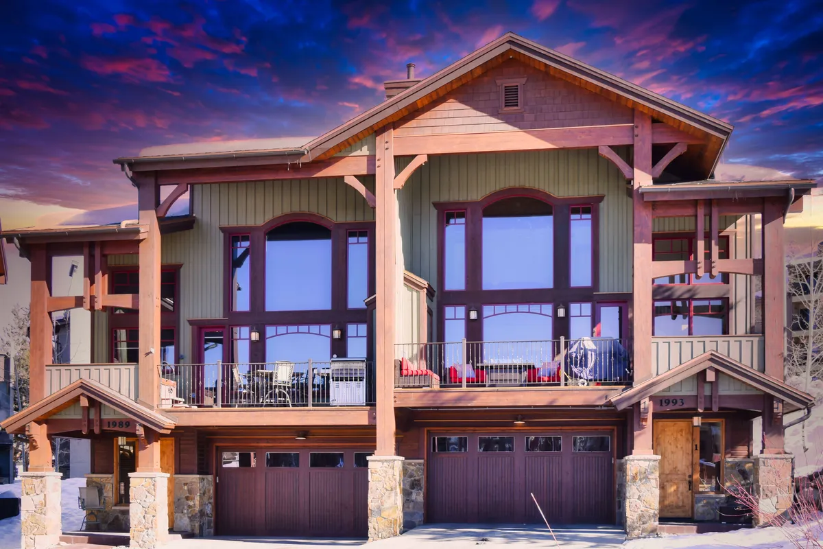 A sophisticated view of a chalet-style exterior with large windows, wooden beams, stone accents, and a balcony featuring comfortable seating. The setting invites guests to enjoy morning coffee or evening stargazing, with the backdrop of mountains and natural beauty.