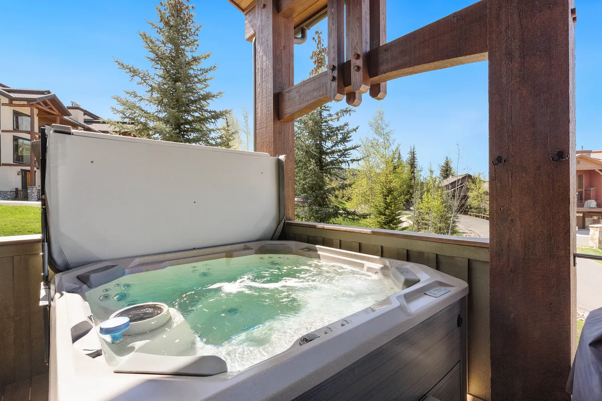 An outdoor deck showcasing a private hot tub surrounded by greenery and mountain views. The tranquil setting invites relaxation, stargazing, and evening gatherings around the firepit just outside the main living spaces.