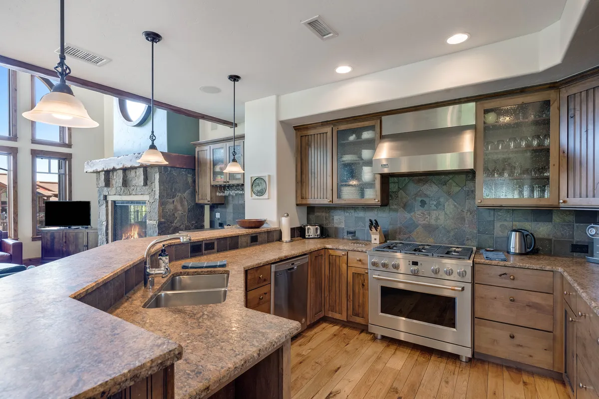 A bright, open kitchen equipped with a large central island, sleek stainless steel appliances, a stone fireplace, and generous windows letting in soft daylight. The layout encourages mingling and group cooking, making it ideal for festive gatherings or casual meals.